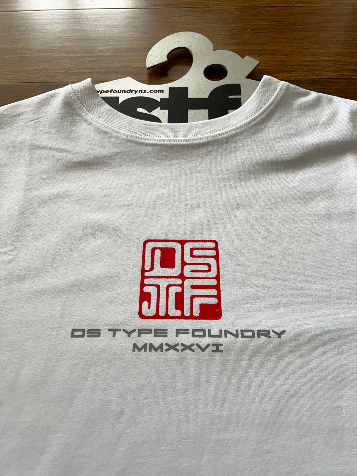 A white crewneck t-shirt laid flat on a wooden surface, featuring a red square logo and the text 'DS TYPE FOUNDRY MMXXVI' printed on the chest. A cardboard hanger with 'dstf' is visible at the collar.