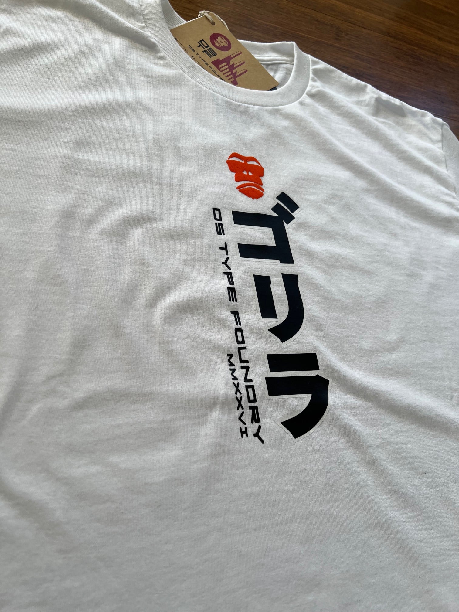 A white crewneck t-shirt laid flat on a dark wood surface, featuring a black stylized logo and the text 'DS TYPE FOUNDRY MMXXVI' printed on the chest, with a brown paper hangtag attached to the collar.